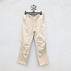Può includere: Pantaloni color cachi appesi a una gruccia nera. I pantaloni hanno un classico design a cinque tasche e ginocchia rinforzate. Realizzati in materiale resistente, adatti all'abbigliamento casual. Lo sfondo è bianco.