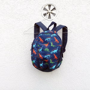 Könnte beinhalten: Ein kleiner blauer Rucksack mit einem Dinosaurier-Muster. Der Rucksack hat einen Reißverschluss und verstellbare Träger.