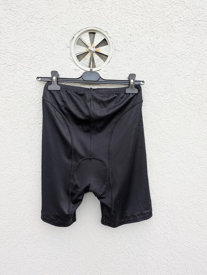 Puede incluir: Pantalones cortos de ciclismo negros confeccionados en tejido sint&eacute;tico liso con costuras visibles a lo largo de las caderas y una zona de la entrepierna reforzada y acolchada. Los pantalones cuelgan de una percha de pl&aacute;stico negra.