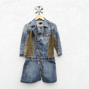 Puede incluir: Un vestido de mezclilla con cremallera frontal y un diseño único. El vestido presenta una combinación de mezclilla azul y paneles verde oliva, creando un contraste elegante. El vestido está colgado en una percha de madera.