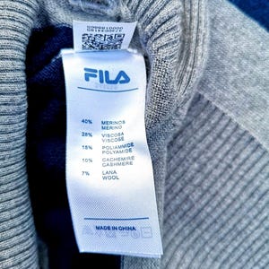 May include: A close-up shot of a grey knit sweater with a Fila label. The label details the composition: 40% Merino wool, 28% viscose, 15% polyamide, 10% cashmere, and 7% wool. The label also states "Made in China".