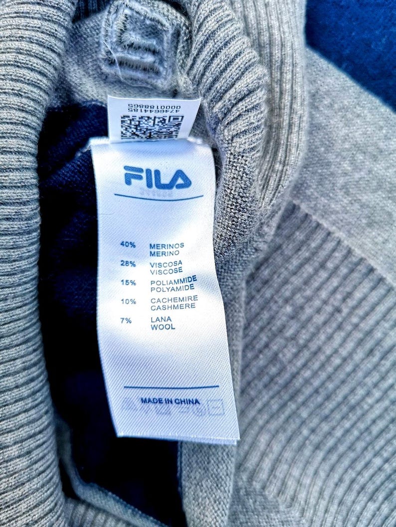 May include: A close-up shot of a grey knit sweater with a Fila label. The label details the composition: 40% Merino wool, 28% viscose, 15% polyamide, 10% cashmere, and 7% wool. The label also states "Made in China".
