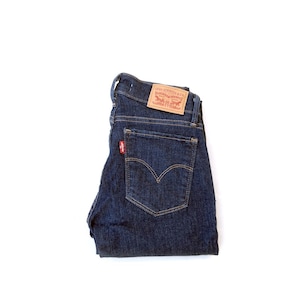 Può includere: Jeans in denim blu scuro con un classico design a cinque tasche. I jeans presentano un'etichetta rossa Levi's sulla tasca posteriore e una toppa in pelle marrone. I jeans sono piegati ordinatamente su sfondo bianco.