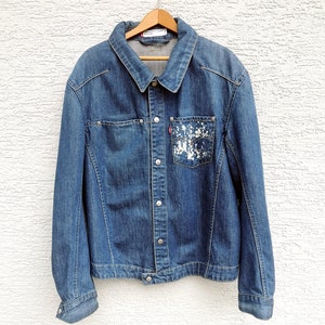 Puede incluir: Chaqueta vaquera azul con cuello clásico y cierre de botones. La chaqueta presenta un bolsillo en el pecho con un diseño de salpicaduras blancas y una etiqueta roja. Cuelga de una percha de madera.