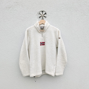 May include: A cream-colored fleece pullover with a high collar and a quarter-zip closure. The pullover features a small Norwegian flag patch on the chest and a small logo on the sleeve. The garment is hanging on a wooden hanger against a white wall.