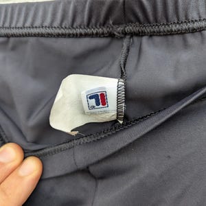 Puede incluir: Pantalones cortos deportivos negros con cintura el&aacute;stica y costuras visibles. Una etiqueta de cuidado blanca est&aacute; unida a la costura interior, mostrando el logotipo de la marca Fila en rojo y azul, junto con texto impreso bajo el logo.