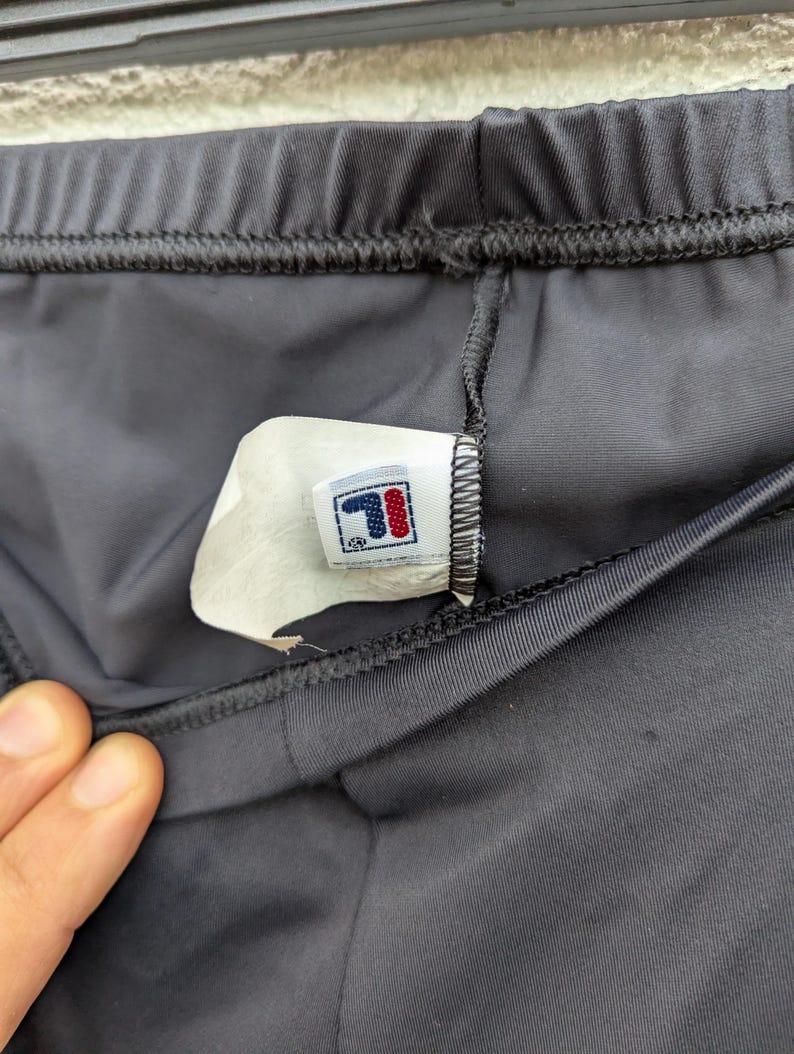 Puede incluir: Pantalones cortos deportivos negros con cintura el&aacute;stica y costuras visibles. Una etiqueta de cuidado blanca est&aacute; unida a la costura interior, mostrando el logotipo de la marca Fila en rojo y azul, junto con texto impreso bajo el logo.