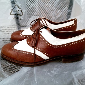 Può includere: Un paio di scarpe in pelle marrone con dettagli in pelle bianca. Le scarpe hanno un design a punta alare e sono allacciate. Le scarpe sono su una superficie di plastica bianca.