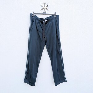 May include: Dark navy blue athletic track pants featuring a white vertical stripe running down each outer leg. The pants have an elastic waistband with a black drawstring, two side pockets, and a small white logo on the upper left thigh area.
