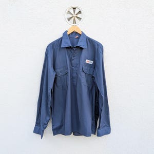 May include: A navy blue long-sleeve shirt with two chest pockets and a small rectangular patch. The shirt is hanging on a wooden hanger against a white wall. The shirt has a button-down collar and buttoned cuffs.