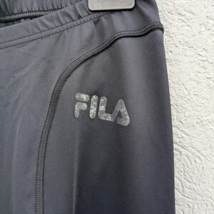 Puede incluir: Leggings deportivos negros con cintura el&aacute;stica alta y un detalle de costura curva a lo largo de la cadera. La palabra FILA est&aacute; impresa en una textura met&aacute;lica plateada desgastada en la zona del muslo de la prenda.