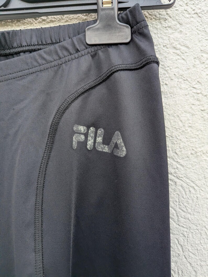 Puede incluir: Leggings deportivos negros con cintura el&aacute;stica alta y un detalle de costura curva a lo largo de la cadera. La palabra FILA est&aacute; impresa en una textura met&aacute;lica plateada desgastada en la zona del muslo de la prenda.