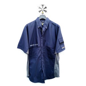 May include: Navy blue short-sleeve button-up shirt with gray side panels. The shirt has a pocket on the left sleeve and the text "MNV-410" printed on the chest. The shirt is hanging on a metal hanger.