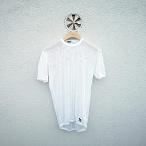 May include: A white, short-sleeved mesh shirt hangs on a wooden hanger against a textured, light gray wall. The shirt has a small, colorful logo near the bottom hem. Above the hanger is a circular vent.