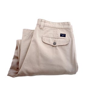 Op de afbeelding: Een beige chino broek gevouwen in de helft. De broek heeft een enkele achterzak met een knoopsluiting en een klein donkerblauw label met de tekst "Dockers" op de rechter achterzak.