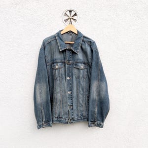Könnte beinhalten: Eine blaue Jeansjacke mit klassischem Design, mit Knopfleiste, Brusttaschen und Kragen. Die Jacke hängt an einem Holzbügel vor einer weißen, strukturierten Wand. Der Denim hat ein verwaschenes Aussehen.