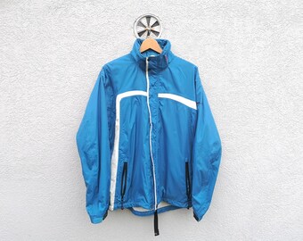 Pode incluir: Uma jaqueta corta-vento azul e branca com fecho de correr. A jaqueta tem detalhes brancos nos ombros e uma faixa branca na frente.