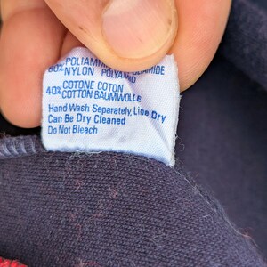 以下が含まれることがあります： 濃紺の生地に縫い付けられた白い洗濯表示タグ。タグには青い文字でナイロン60%、コットン40%と記載されており、手洗い、陰干し、ドライクリーニング、漂白剤使用不可の指示が記されています。