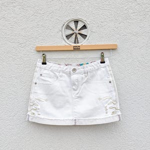 May include: A white denim mini skirt featuring a five-pocket design, a button and zip fly closure, and light-coloured embroidered wave-like patterns on the front panels. The hem is finished with a raw, frayed edge and a rolled cuff detail.