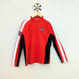Könnte beinhalten: Rotes Langarmshirt mit schwarzen Seitenteilen und weißen Streifen an den Ärmeln. Mit einem kleinen Logo-Patch auf der Brust und am Ärmel. Das Shirt hängt an einem Holzbügel.