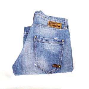 Op de afbeelding: Een paar blauwe denim jeans gevouwen in de helft, met de achterzak zichtbaar en een bruin leren label met de tekst "The Authentic Jeans" en "Melvin & Hamilton".