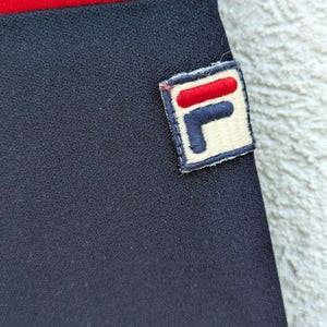 以下が含まれることがあります： 上端に赤い水平の帯がある黒い布製の衣類。白い背景に赤い水平のバーと紺色の文字Fで構成されたFilaロゴの長方形のワッペンが、黒い部分に縫い付けられています。