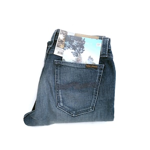 May include: A pair of blue denim jeans with a back pocket that has a small brown leather patch with the text "Nudie Jeans" on it. A folded piece of paper with a blue and white design is tucked into the back pocket.