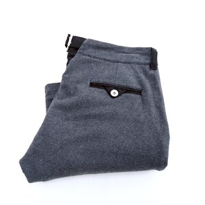 May include: Folded pair of gray trousers with a black waistband and pocket accents. The pocket features a black trim and a white button. The pants are made of a soft, textured fabric.