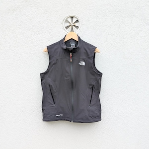 May include: A charcoal gray vest with a full-length zipper and two side pockets. The vest has the North Face logo on the chest and is hanging on a wooden hanger against a white wall.