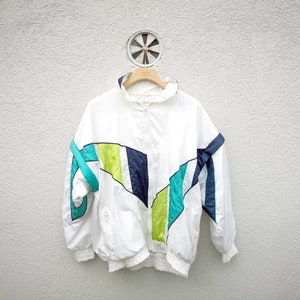 May include: White vintage windbreaker jacket with a high collar and a zipper closure. The jacket features a color block design with teal, navy, and lime green accents. The jacket is hanging on a wooden hanger against a white wall.