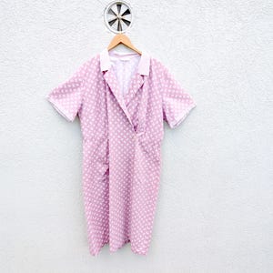 May include: A short-sleeved wrap dress in a light pink colour featuring a repeating white floral pattern. The dress has a solid white collar, a V-neckline, and a wrap-style front closure that secures at the waist, creating a relaxed, knee-length silhouette.