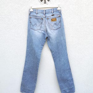 May include: Light blue denim jeans with a brown leather patch on the back pocket. The jeans are hanging on a white hanger against a white wall.
