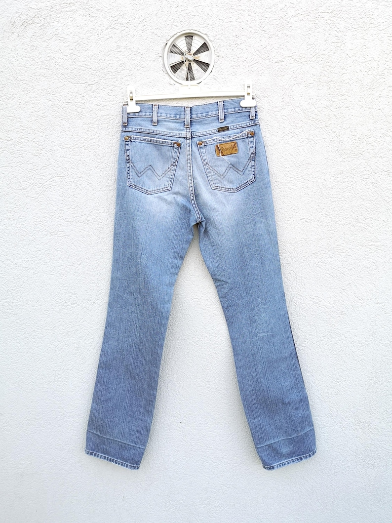 May include: Light blue denim jeans with a brown leather patch on the back pocket. The jeans are hanging on a white hanger against a white wall.
