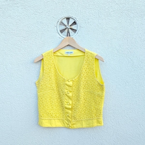 May include: A bright yellow sleeveless vest featuring a textured, floral-patterned lace overlay on the front panels. The garment has a scoop neckline, a button-down front with four matching yellow buttons, and a solid yellow hem and back.