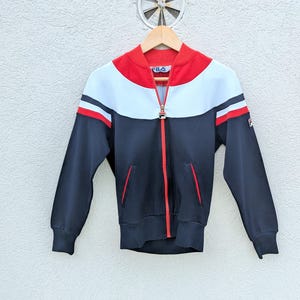 以下が含まれることがあります： ネイビーブルー、ホワイト、レッドのカラーブロックデザインが特徴のFilaジップアップトラックジャケット。リブ編みの襟、袖口、裾、フルレングスの赤いジッパー、2つのサイドポケット、胸と左袖にFilaのロゴがあしらわれています。