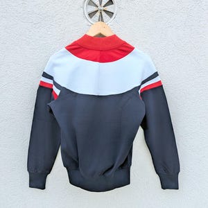 以下が含まれることがあります： 赤い襟、水色の胸パネル、ダークネイビーのボディというカラーブロックデザインの長袖トラックジャケット。袖には肩の縫い目に沿って白と赤の水平ストライプがあしらわれており、全体的にスポーティーな印象を与えるデザインとなっている。