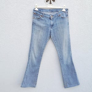 May include: Light blue denim jeans with a bootcut leg opening. The jeans are hanging on a white hanger against a white wall.