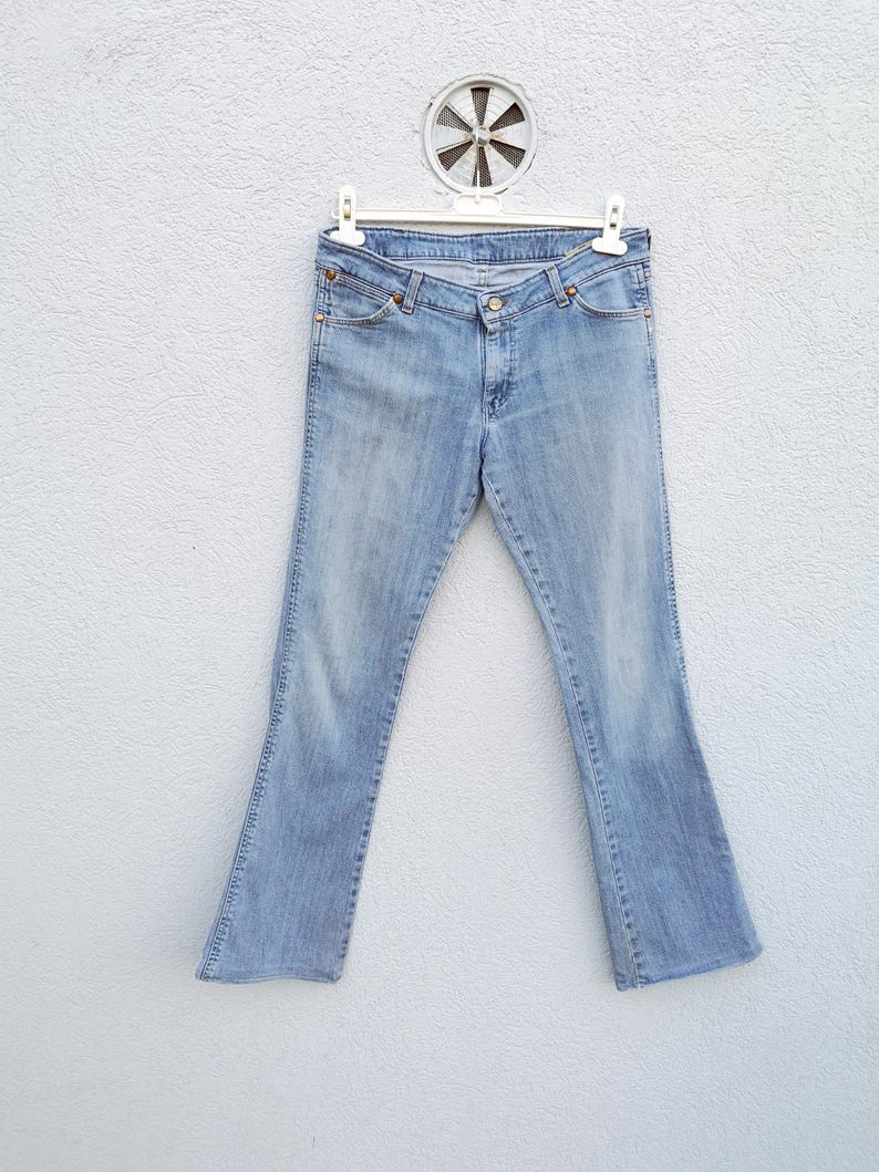 May include: Light blue denim jeans with a bootcut leg opening. The jeans are hanging on a white hanger against a white wall.