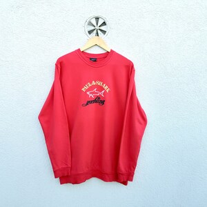 May include: Red long-sleeve sweatshirt with a white embroidered shark and the text "Paul & Shark" and "yachting" in black.