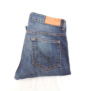 Puede incluir: Vaqueros de mezclilla azul doblados con un parche de cuero marrón en la cintura trasera. Los vaqueros tienen un diseño clásico de cinco bolsillos, sobre fondo blanco.