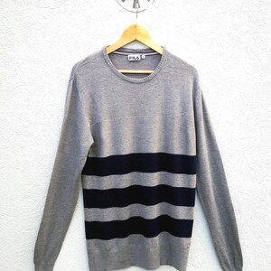 May include: A grey crew neck jumper with navy blue horizontal stripes. The jumper is hanging on a wooden hanger against a white wall. The brand name "FILA" is visible on the collar.