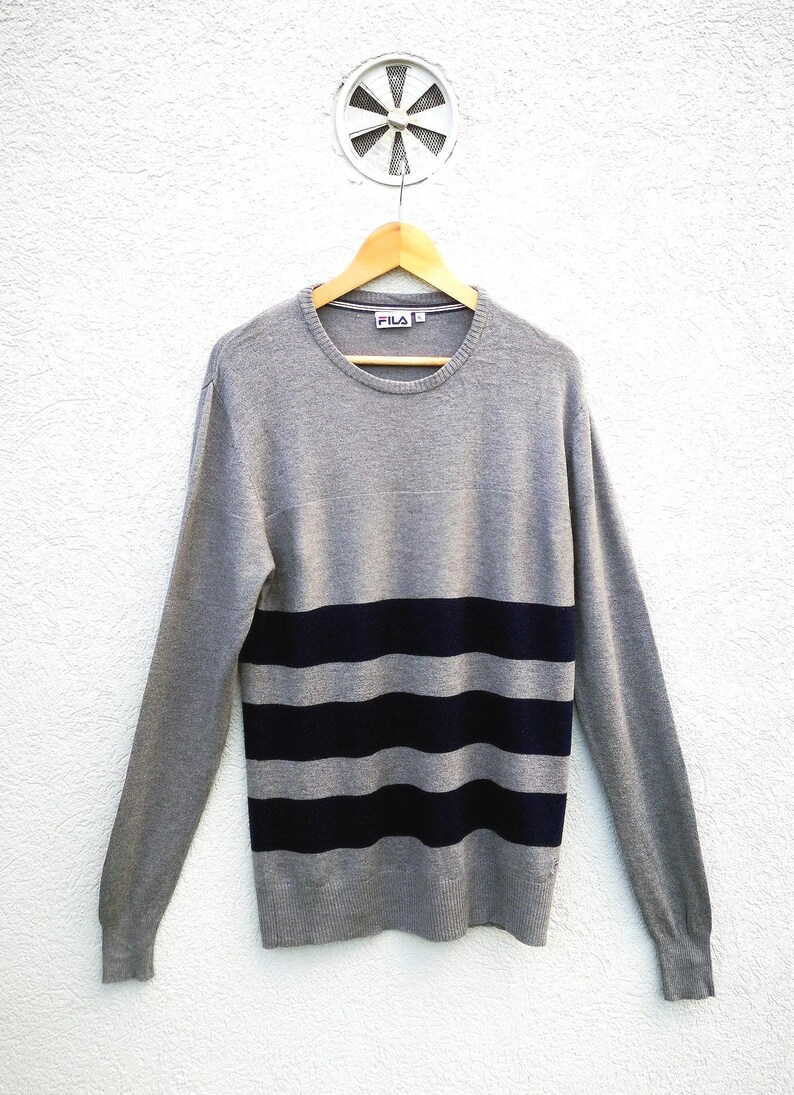 May include: A grey crew neck jumper with navy blue horizontal stripes. The jumper is hanging on a wooden hanger against a white wall. The brand name "FILA" is visible on the collar.
