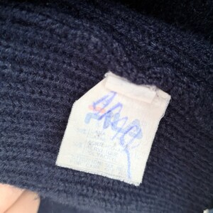 Puede incluir: Primer plano de una tela de punto azul marino con una etiqueta blanca. La etiqueta tiene texto y un logotipo en azul y rojo. La tela parece ser parte de un su&eacute;ter o prenda similar.