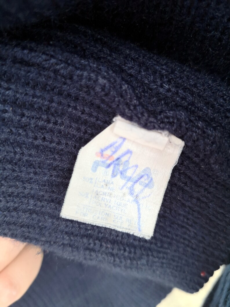 Puede incluir: Primer plano de una tela de punto azul marino con una etiqueta blanca. La etiqueta tiene texto y un logotipo en azul y rojo. La tela parece ser parte de un su&eacute;ter o prenda similar.