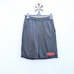 Puede incluir: Pantalones cortos deportivos de color gris oscuro con una textura de malla, colgados de una percha negra. Los pantalones cortos tienen bolsillos y un parche rectangular rojo con la palabra "PYREX" en letras blancas. Los pantalones cortos se muestran sobre un fondo blanco.