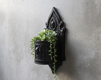 May include: Black wall-mounted planter with a gothic design, featuring a skull and ornate details. The planter holds green succulents with trailing stems, creating a striking contrast against the dark fixture. A unique piece for home decor.