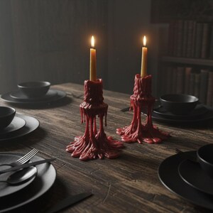 May include: A dimly lit dining table set for a meal. Two lit candles in red, dripped-wax holders are the focal point. Black plates, bowls, and silverware are arranged on the wooden table. A bookshelf is in the background.
