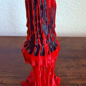 May include: A 3D-printed, abstract candle holder in red and black. The design features a cylindrical black core with red, dripped-wax-like accents. The base is a flared, melted-wax design. The candle holder is displayed on a wooden surface.