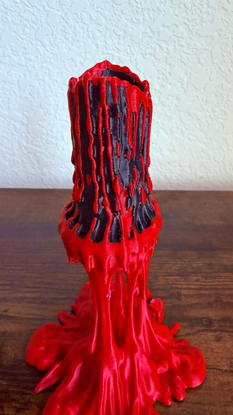 May include: A 3D-printed, abstract candle holder in red and black. The design features a cylindrical black core with red, dripped-wax-like accents. The base is a flared, melted-wax design. The candle holder is displayed on a wooden surface.
