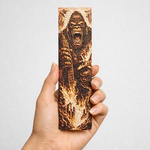 May include: A rectangular wooden bookmark featuring a detailed illustration of a roaring gorilla holding a building, with a burning city and airplanes. The artwork is in shades of brown and orange.